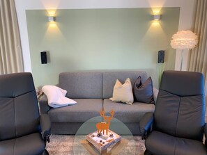 Una sala de estar con un sofá gris, dos sillones negros, una mesa de centro con un objeto decorativo e iluminación en la pared.