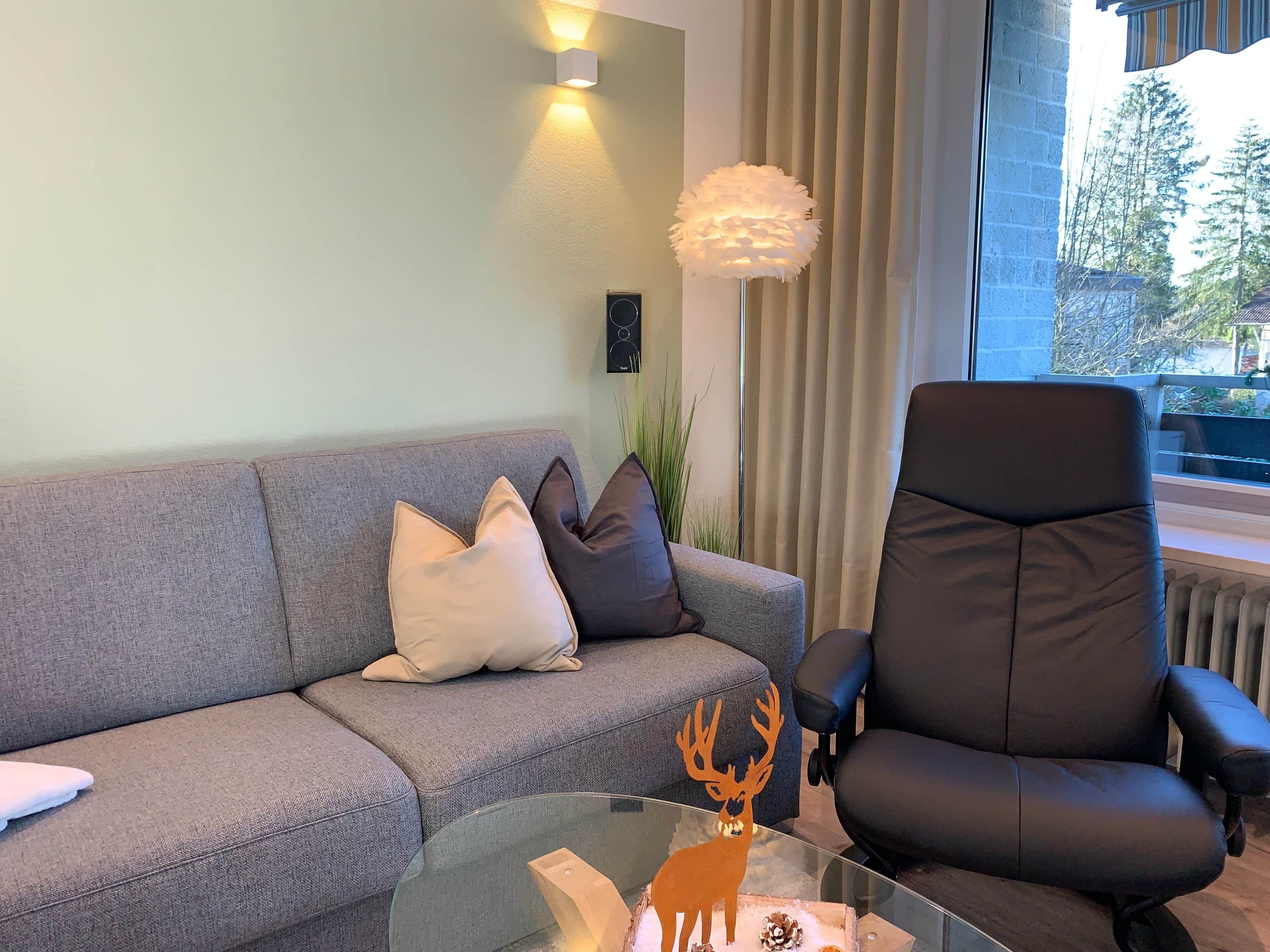 Un salon moderne avec un canapé gris, un fauteuil en cuir noir, une table basse en verre ornée d’un motif de cerf et une grande fenêtre avec des rideaux.