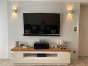 Ein Flachbildfernseher an der Wand, zwei Wandleuchten, ein Holztischchen mit einer Uhr, Fernbedienungen und einem Lautsprecher.