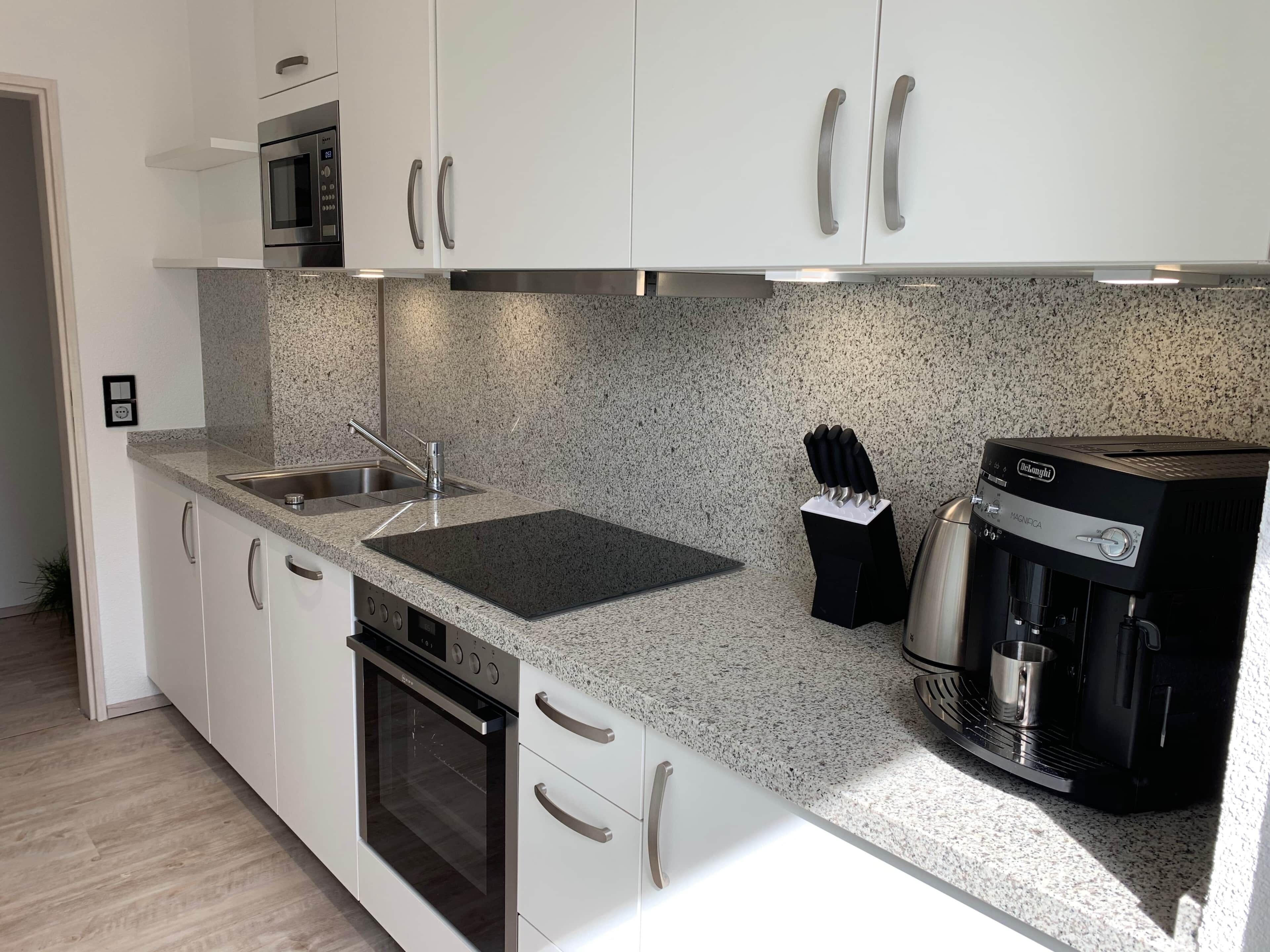Une cuisine moderne avec des meubles blancs, un plan de travail en granit, un four encastré, une machine à café et un four à micro-ondes.