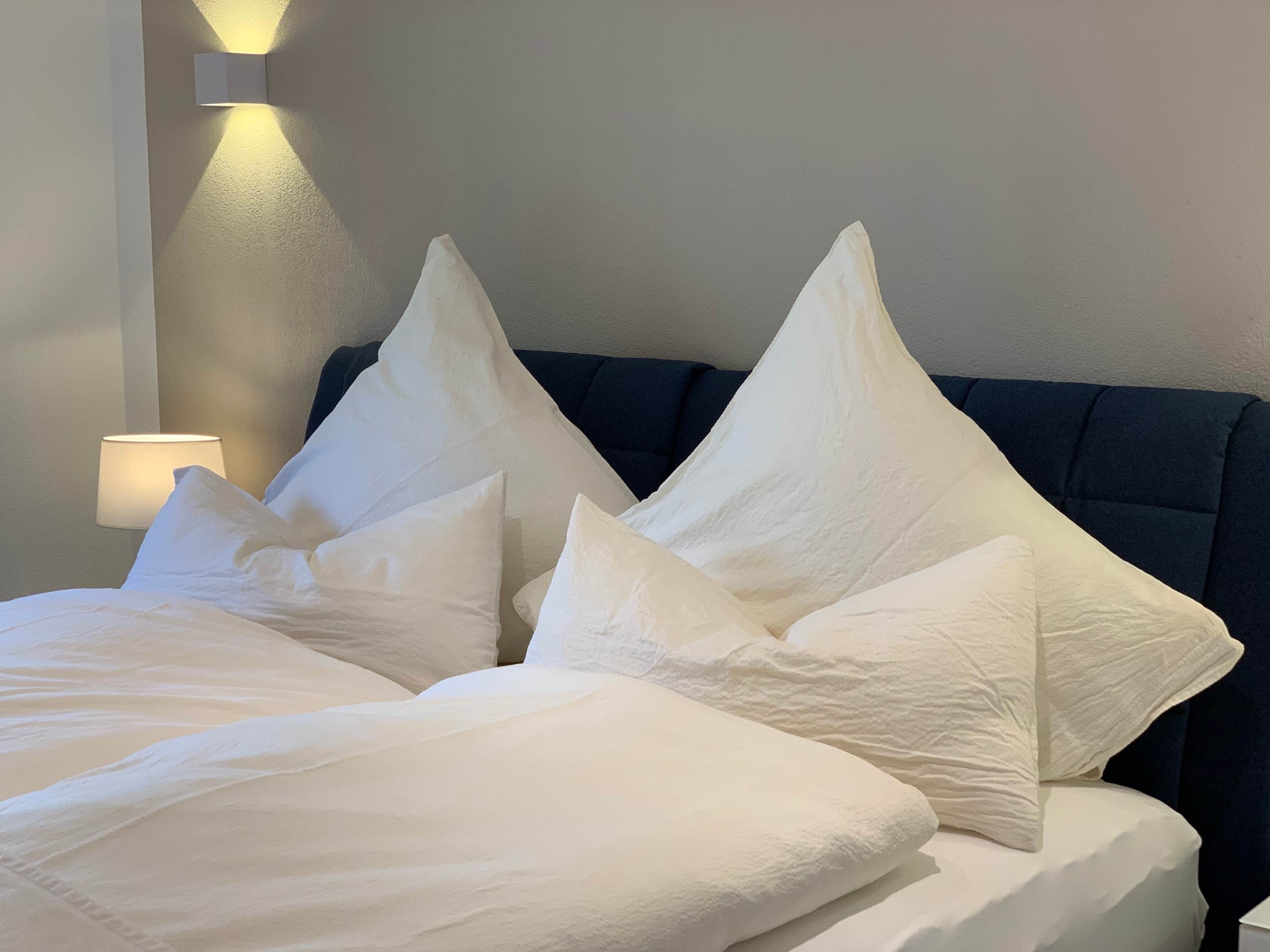 Cama com roupa de cama branca e várias almofadas, cabeceira escura e luminária fixada na parede.