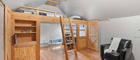 A loft bedroom with a wooden ladder, a black leather chair, and a view of the sky.