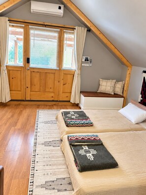 A room with a sloped ceiling, wooden door, air conditioning unit, two beds with patterned blankets, a bench, and a window with curtains. - Historic Old Town Attic Loft—Steps from Albuquerque’s Best Sights (Albuquerque)