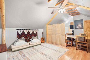 Una camera da letto accogliente in mansarda, con travi a vista, una ventola a soffitto, un letto con cuscini, una chitarra, un tavolo da pranzo con sedie e un'ampia finestra.