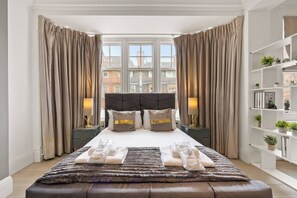 Une chambre d’hôtel avec un grand lit, deux tables de chevet avec des lampes, une vue sur les immeubles à travers de grandes fenêtres et une bibliothèque agrémentée de plantes.