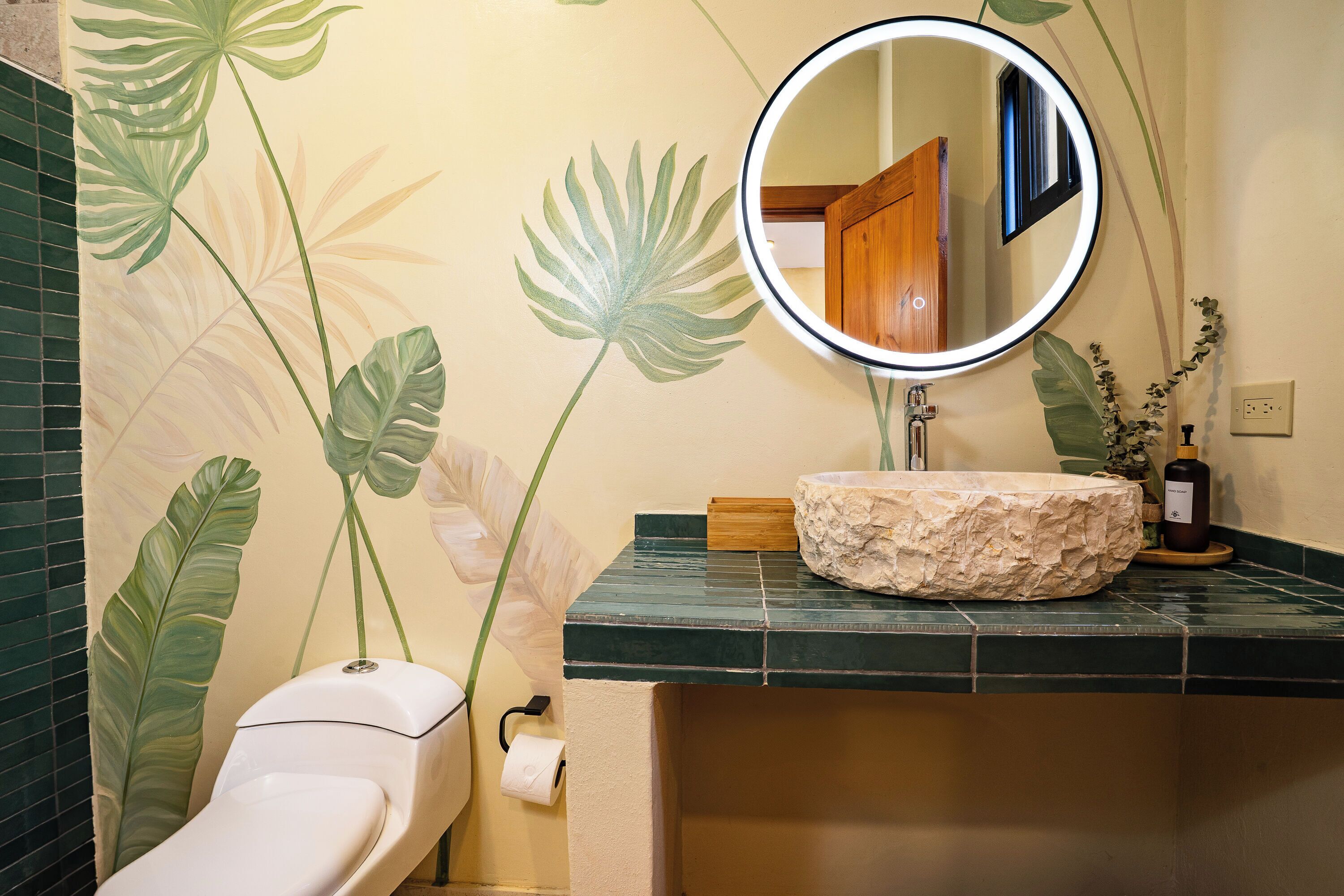 A bathroom with a toilet, a sink with a stone basin, a mirror, and a wall with a tropical leaf mural.