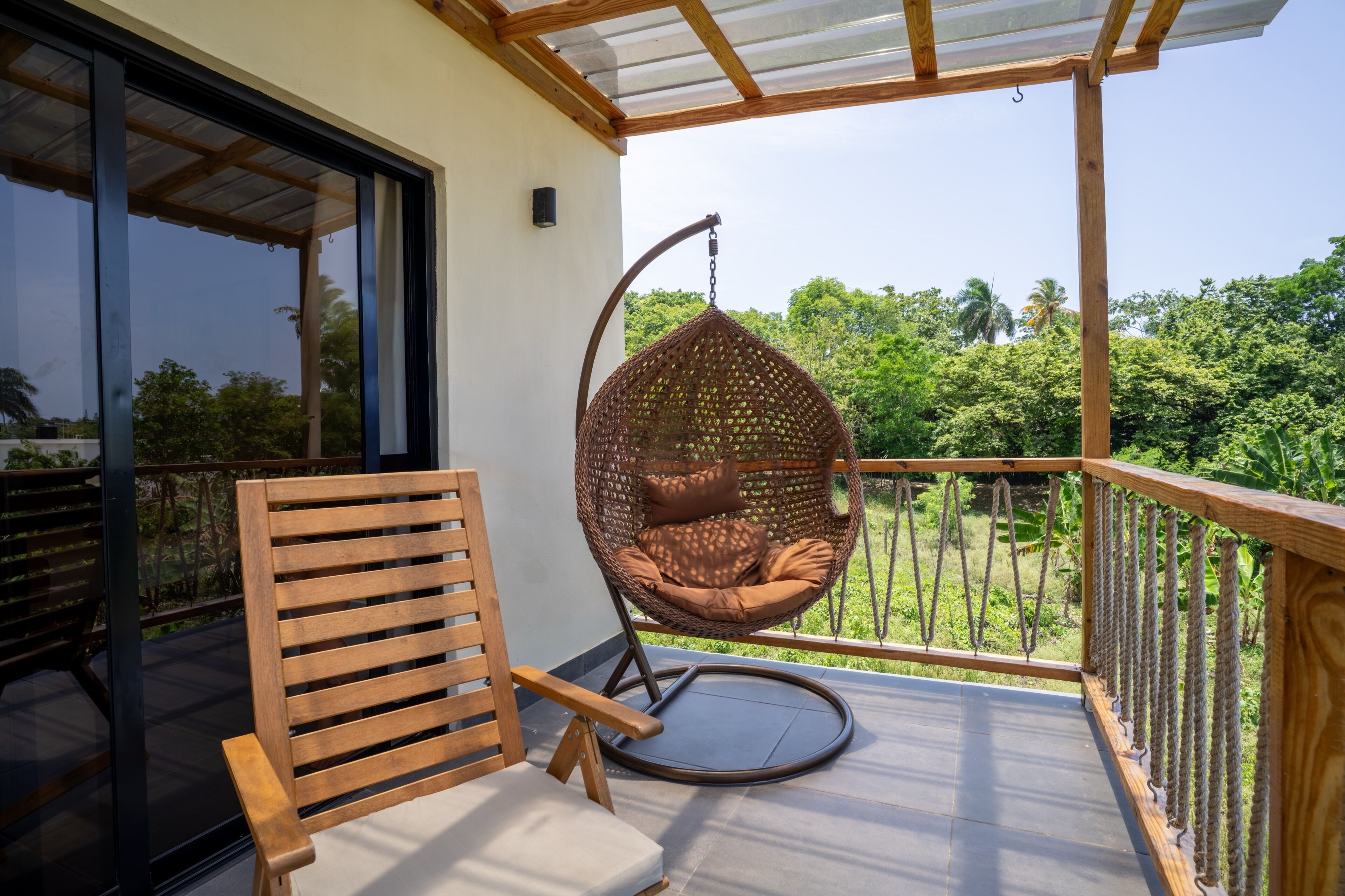 Un balcón con una silla de madera, una mecedora de ratán colgante y vistas de vegetación exuberante.