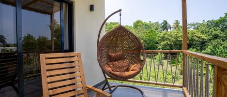 Un balcón con una silla de madera, una mecedora de ratán colgante y vistas de vegetación exuberante.