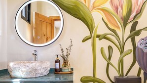 A bathroom with a round mirror, a sink, a decorative wall painting, a trash bin, and a woven basket.