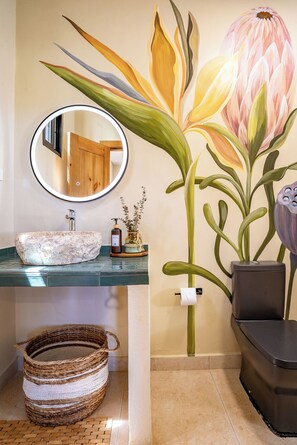 A bathroom with a round mirror, a sink, a decorative wall painting, a trash bin, and a woven basket.