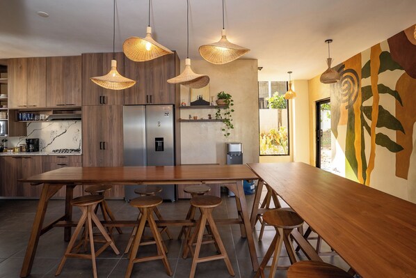 A modern kitchen with wooden cabinets, a stainless steel refrigerator, and a large wooden dining table with stools.