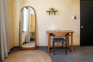A room with a wooden table, a chair, a mirror, a small shelf with a plant, and a door.