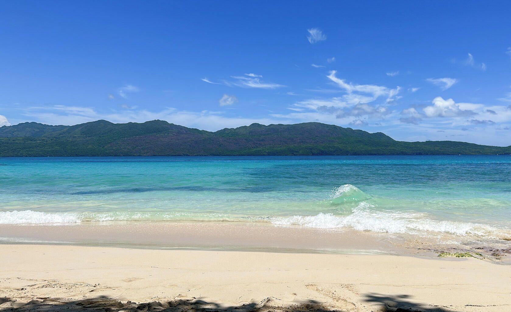 Ein Strand mit klarem türkisblauem Wasser, einem sandigen Ufer und einer Bergkette im Hintergrund.