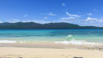 Una playa con agua turquesa cristalina, una orilla arenosa y una cadena montañosa al fondo.