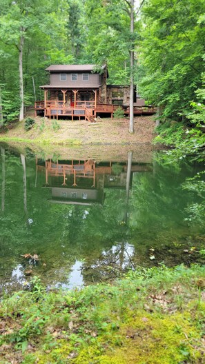 Una cabaña con terraza que da a un lago, rodeada de un bosque denso.