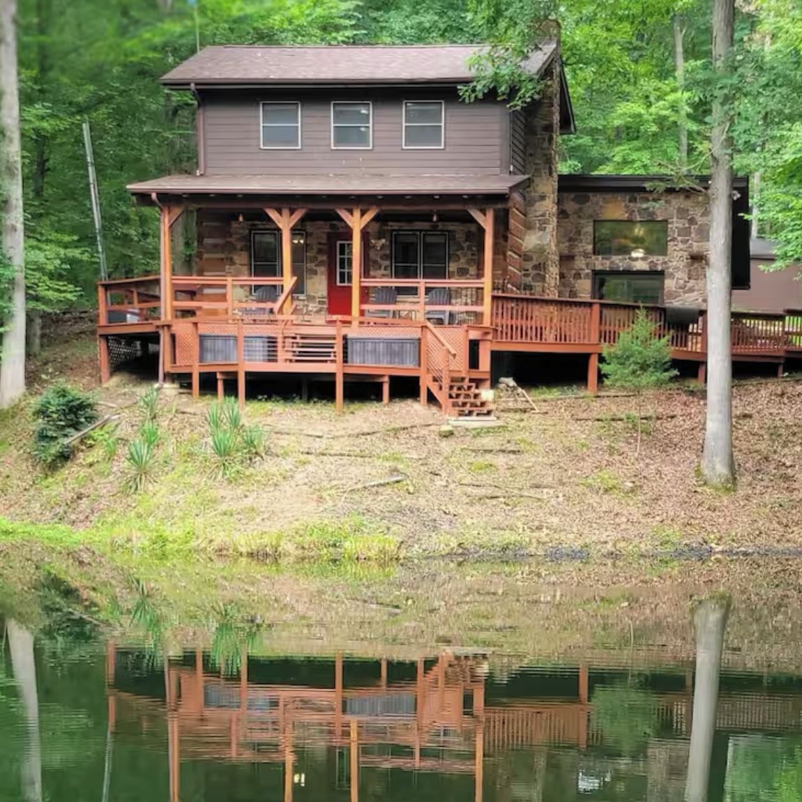 A two-story cabin with a wooden deck and stone walls, situated next to a body of water.