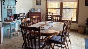 Un coin repas avec une table en bois, des chaises, un ventilateur de plafond et une fenêtre donnant sur les arbres.