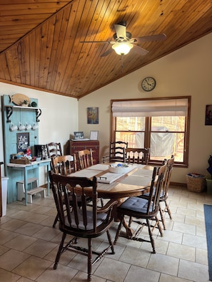 Un comedor con una mesa de madera, sillas, un ventilador de techo y una ventana con vistas a árboles.