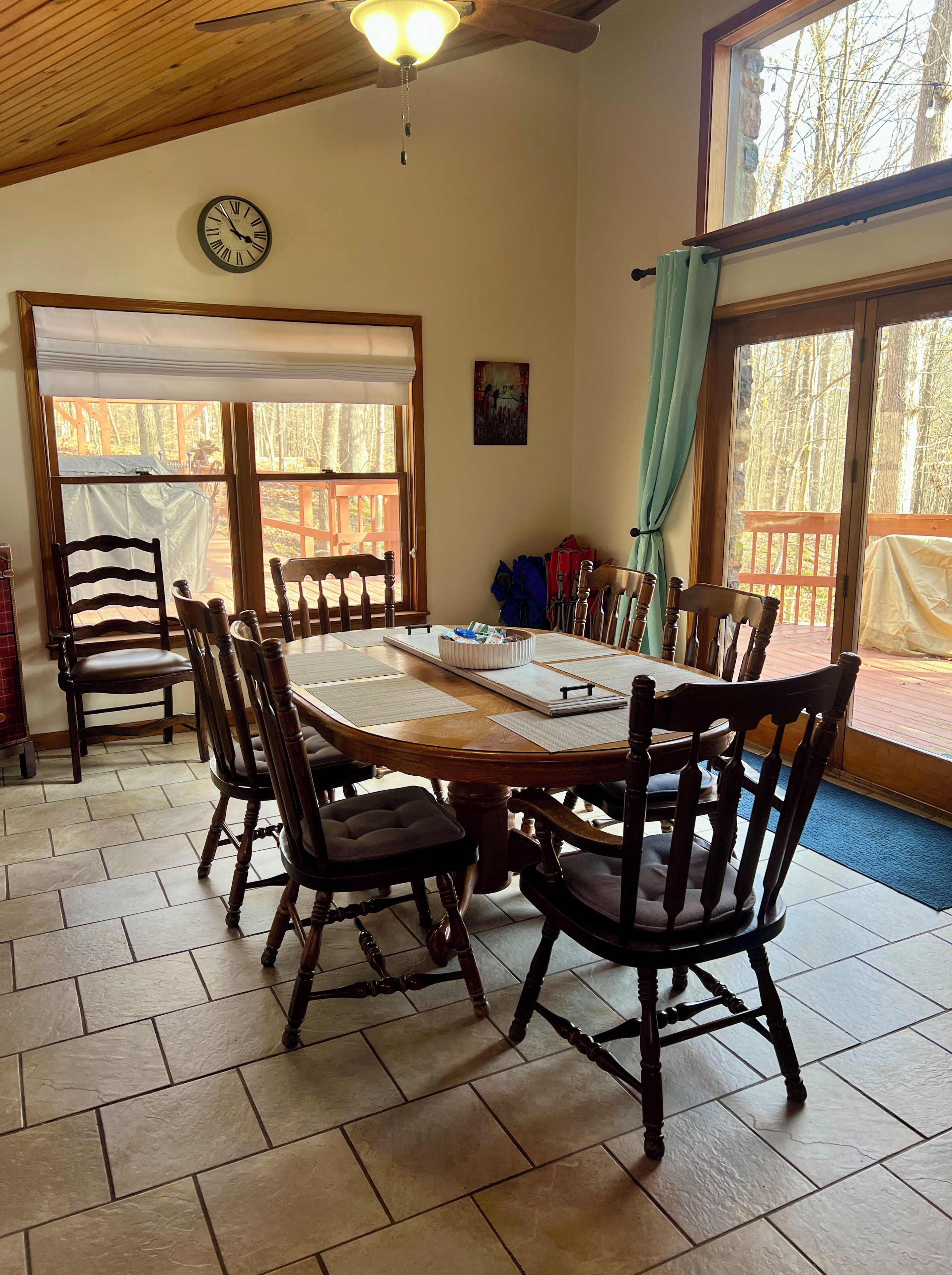A dining area with a wooden table and chairs, a clock on the wall, and windows with curtains.