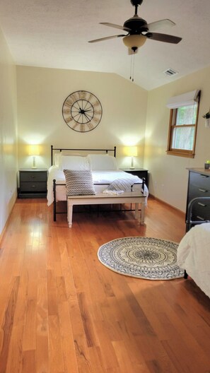 Un dormitorio con cama, lámparas de noche, ventilador de techo, un reloj de pared y una ventana con cortina.