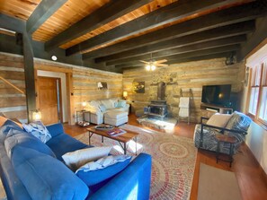 L’intérieur d’une cabane en rondins, avec un canapé, un fauteuil, une télévision, une cheminée et des poutres en bois au plafond.
