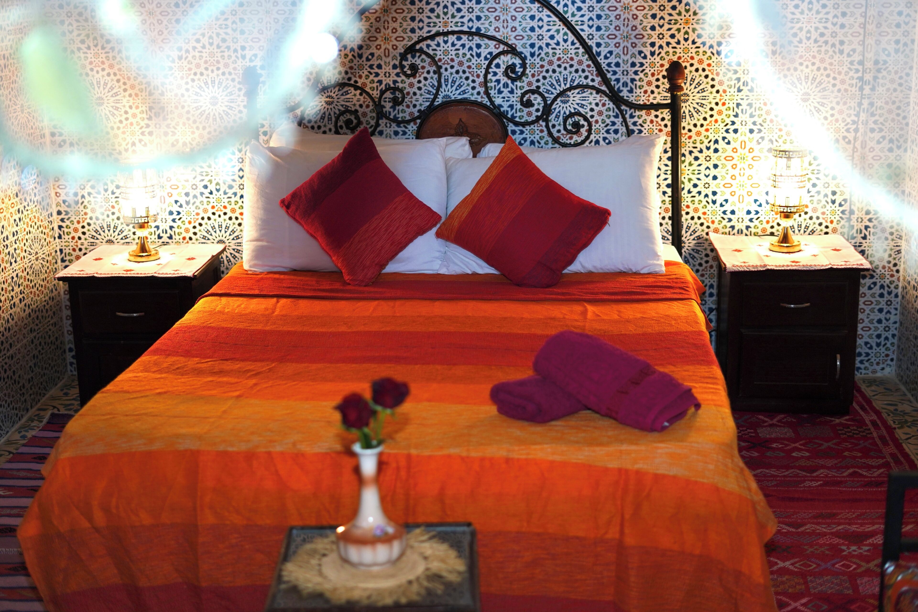 A bed with a red and orange bedspread, two red pillows, a vase with flowers, and a purple towel on the floor.