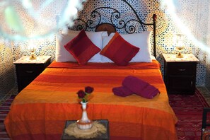 A bed with a red and orange bedspread, two red pillows, a vase with flowers, and a purple towel on the floor. - Riad Dar Ceramic: Heart of Medina w/ AC, WiFi & TV (Marrakesh)