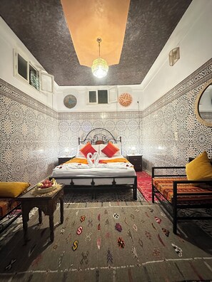 Une chambre avec un lit, deux tables de chevet, une table avec des fruits et un tapis à motifs.