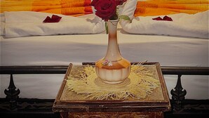 Un llit amb un capçal decoratiu, un vas amb una flor i un llençol amb un dibuix.