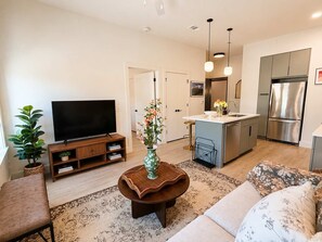 A modern living room with a flat-screen TV, a coffee table with a vase, a sofa, and a kitchen area with a refrigerator and a sink. - The Jardin Rose Suite | Near Downtown (Savannah)