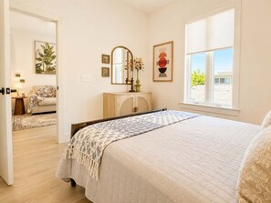 Quarto com cama, penteadeira, espelho, janela e porta que dá para outro cômodo.