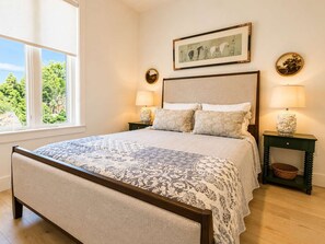 A bedroom with a large bed, two bedside tables with lamps, a framed picture on the wall, and a window with a view of trees. - The Jardin Rose Suite | Near Downtown (Savannah)