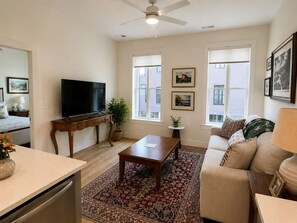 A living room with a flat-screen TV, a coffee table, a sofa, a dining table, and a ceiling fan. - The Jardin Lily Suite | Near Downtown (Savannah)