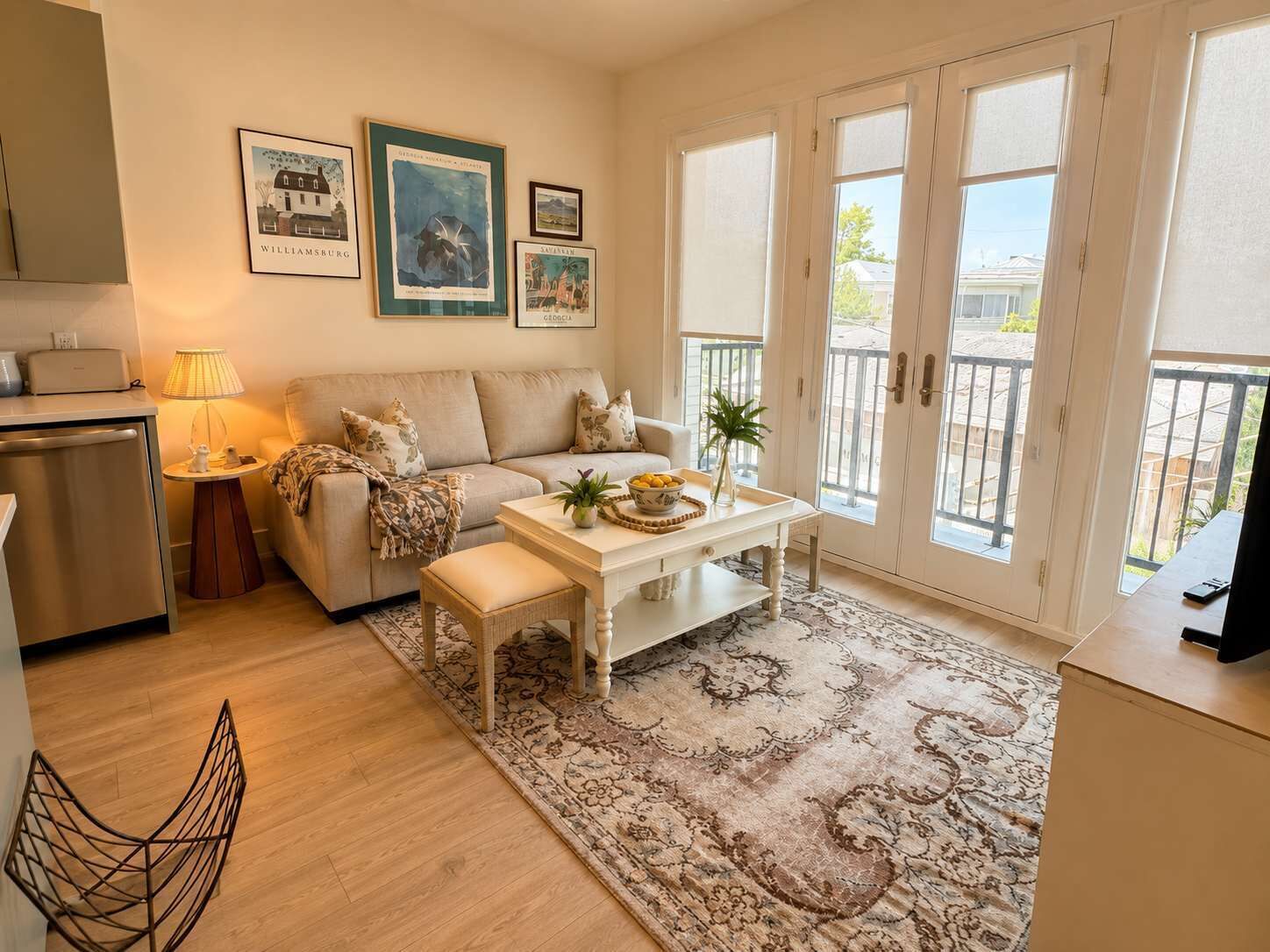 A living room with a sofa, a coffee table, a lamp, and a dining table. There are framed pictures on the wall and a balcony with a railing.