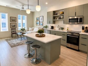 A modern kitchen with a central island, bar stools, a microwave, oven, and a dining area with a sofa and coffee table. - The Jardin Iris Suite | Near Downtown (Savannah)