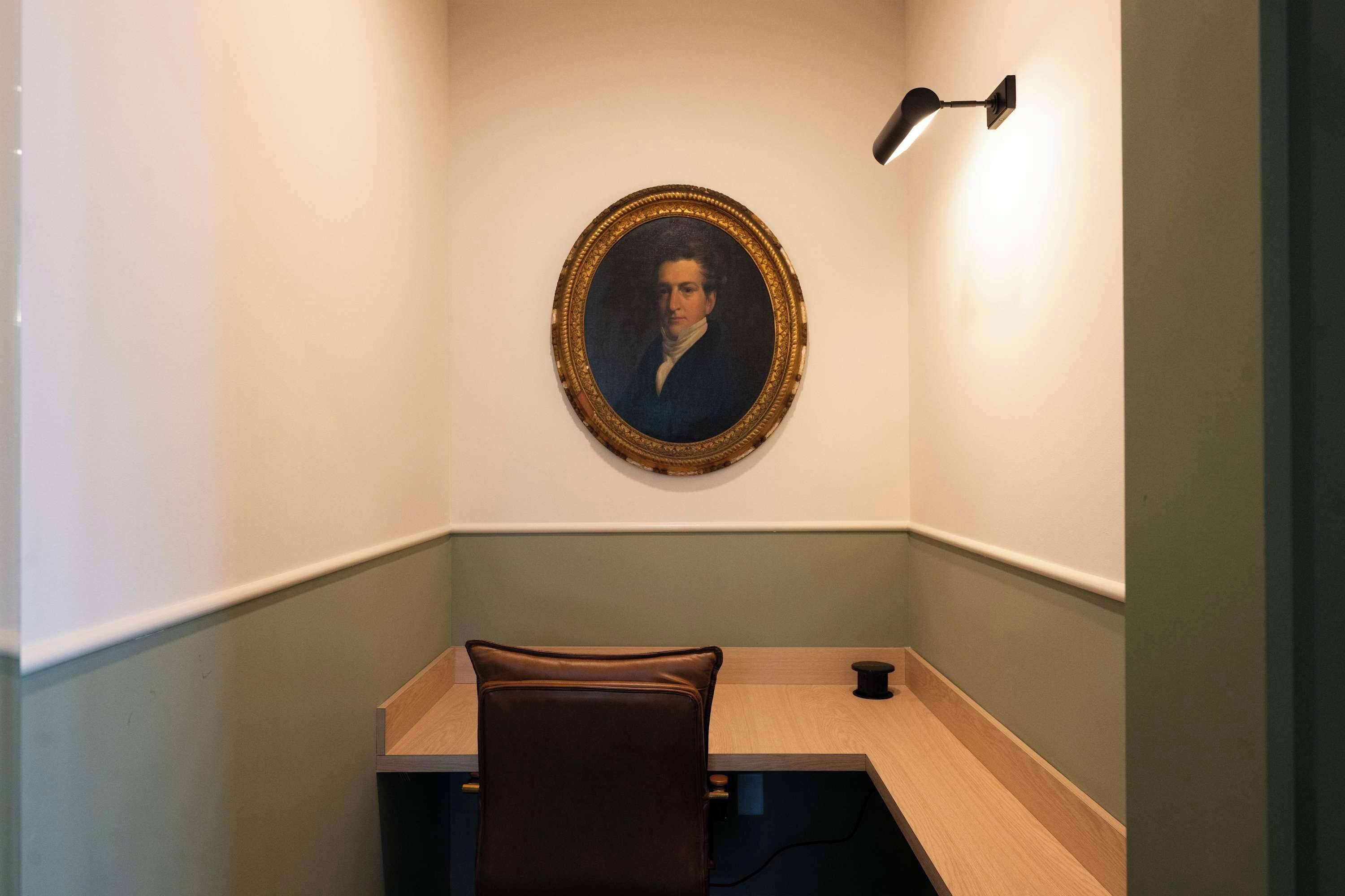 Una habitación con escritorio, una silla, un retrato enmarcado en la pared y una lámpara de pared.