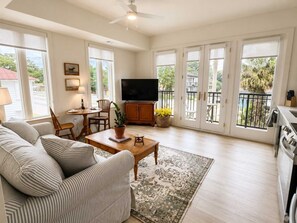 A spacious living room with a sofa, a coffee table, a television, and a balcony with a view. - The Jardin Daisy Suite | Near Downtown (Savannah)