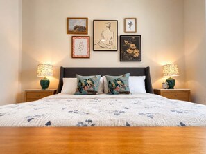 A bedroom with a large bed, two bedside tables with lamps, and four framed artworks on the wall. - The Jardin Daisy Suite | Near Downtown (Savannah)
