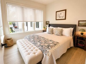 A bedroom with a large bed, a bedside table with a lamp, a window with blinds, and a wall-mounted picture. - The Jardin Briar Suite | Near Downtown (Savannah)
