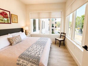 A bedroom with a large bed, a chair, a nightstand, a lamp, and a window with a view of buildings outside. - The Jardin Elm Suite | Near Downtown (Savannah)