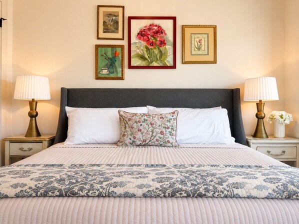 Een net opgemaakt bed met een bloemenprint deken en kussens, aan weerszijden van twee nachtkastjes met lampen, en een wand met ingelijste kunstwerken.