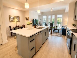 A modern kitchen with a central island, a dining area with a table and chairs, and a living space with a sofa and a coffee table. - The Jardin Elm Suite | Near Downtown (Savannah)
