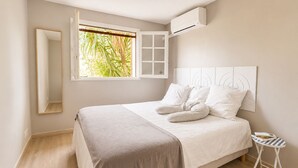 Et soveværelse med seng, et vindue med udsigt til grøn bevoksning, et sengebord og en aircondition.