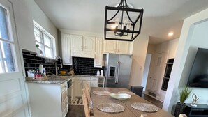 Uma cozinha moderna com armários brancos, uma geladeira de aço inoxidável e uma área de jantar com mesa e cadeiras de madeira.