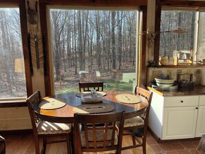 Ein Essbereich mit Tisch und Stühlen, Blick auf einen Wald durch große Fenster, sowie eine Küchenzeile mit Schränken und einer Arbeitsplatte.