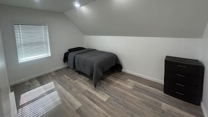 Ein Schlafzimmer mit Bett, Kommode, Fenster mit Jalousien und Einbauleuchten.