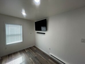 Una habitació amb una finestra gran, televisió plana penjada a la paret i terra de fusta.
