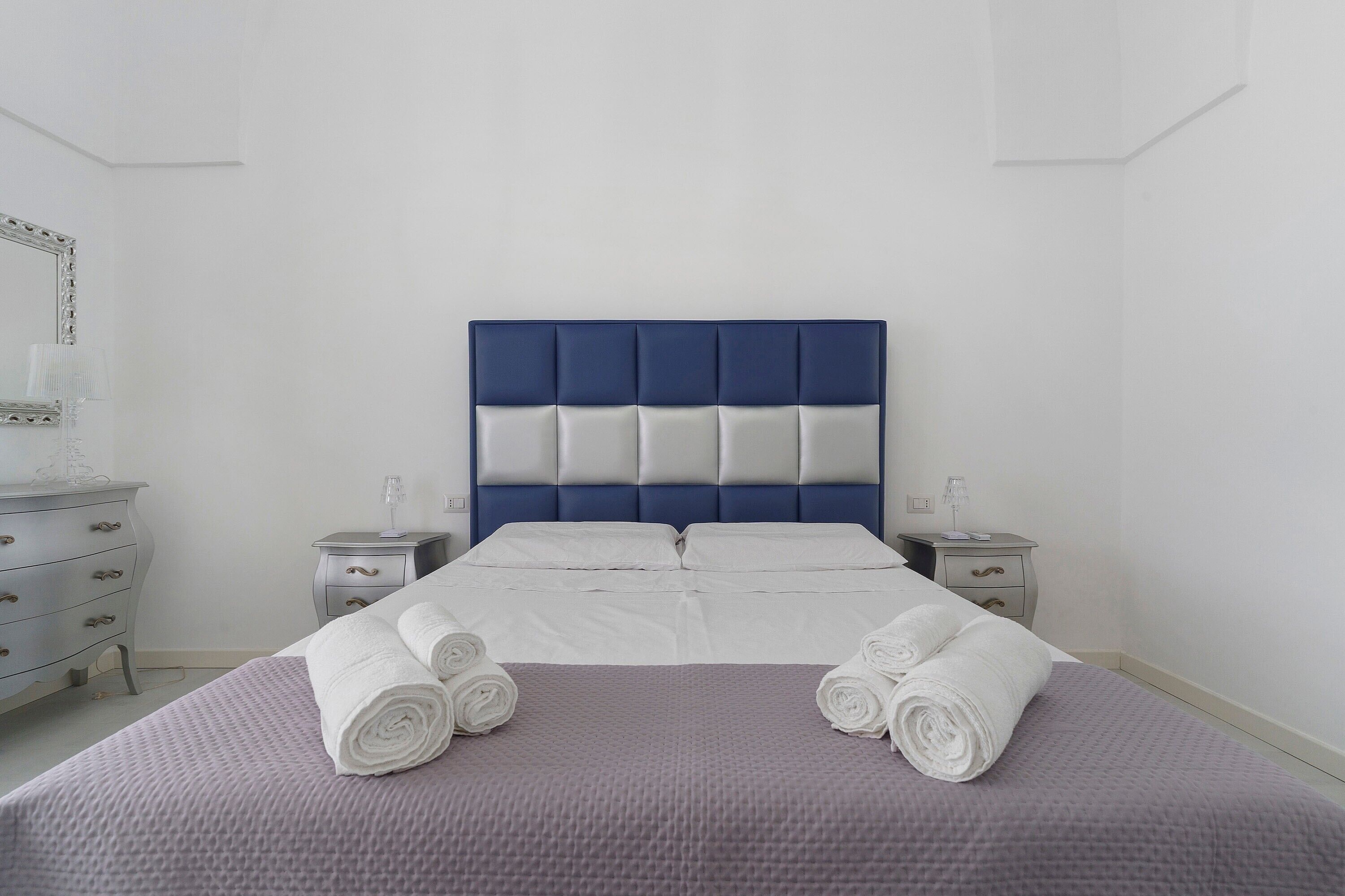 Ein ordentlich bezogenes Bett mit blauem Kopfteil, zwei Nachttischen und einer Kommode mit Schubladen.