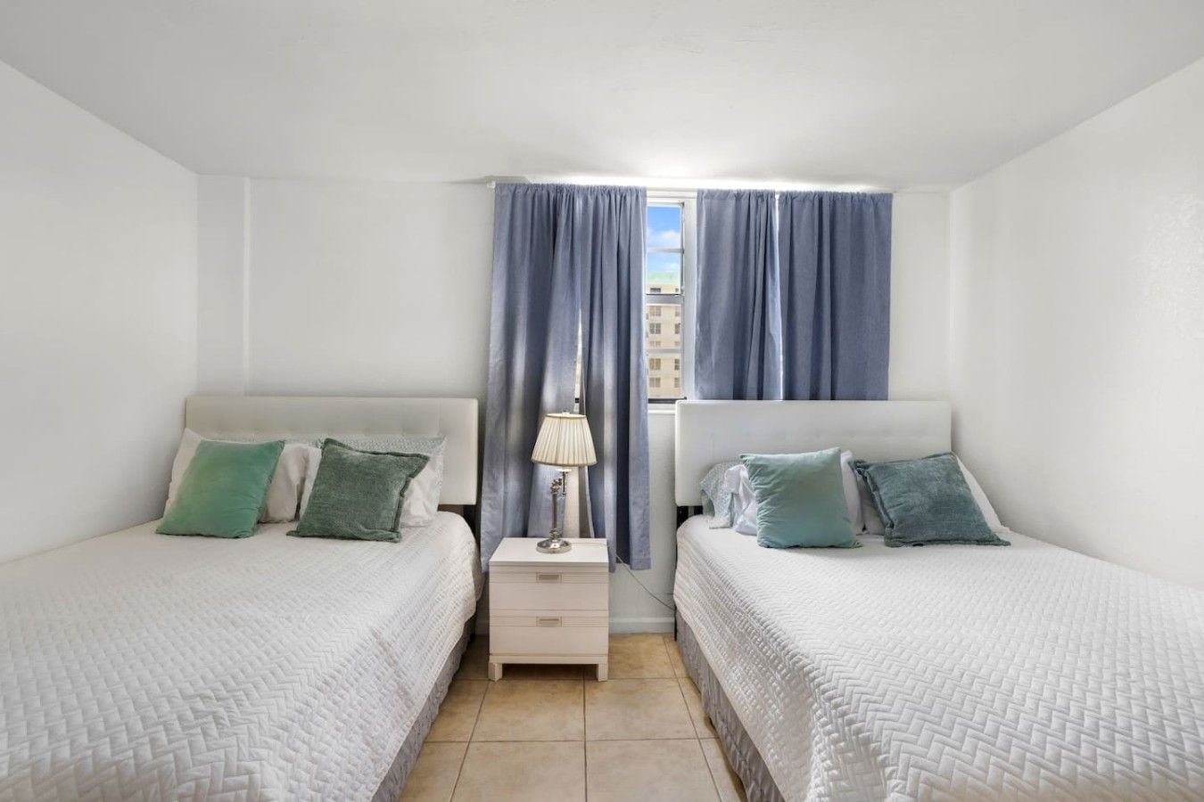 A twin room with two beds, a nightstand, a lamp, and a window with curtains.