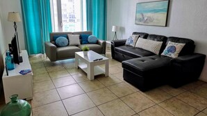 A living room with a sofa, a coffee table, and a TV. There is a window with curtains, a lamp, and a painting on the wall. - Vive la experciencia frente al mar (Hollywood)